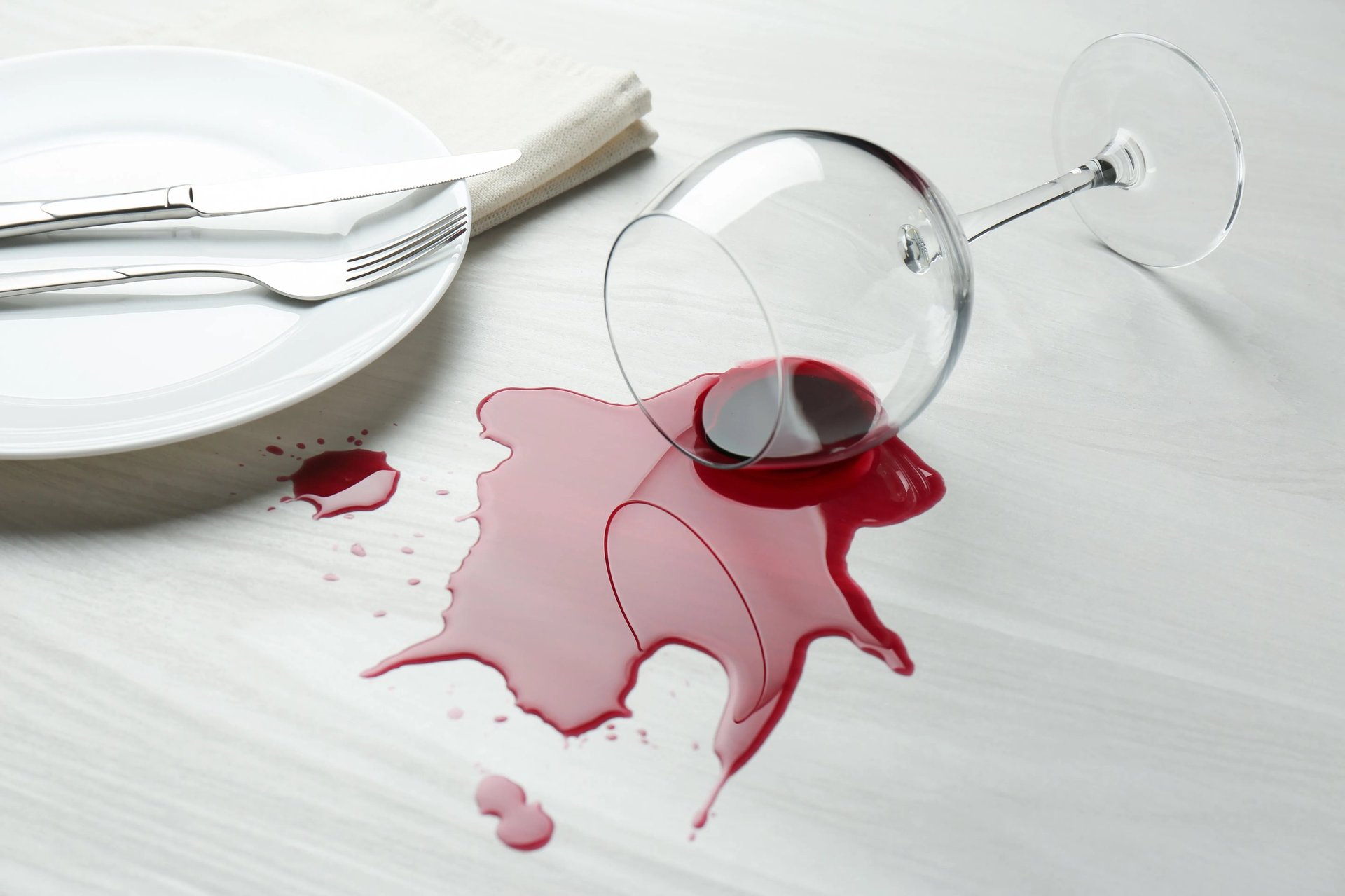 Overturned wine glass spilling red wine onto white surface with plate and fork nearby
